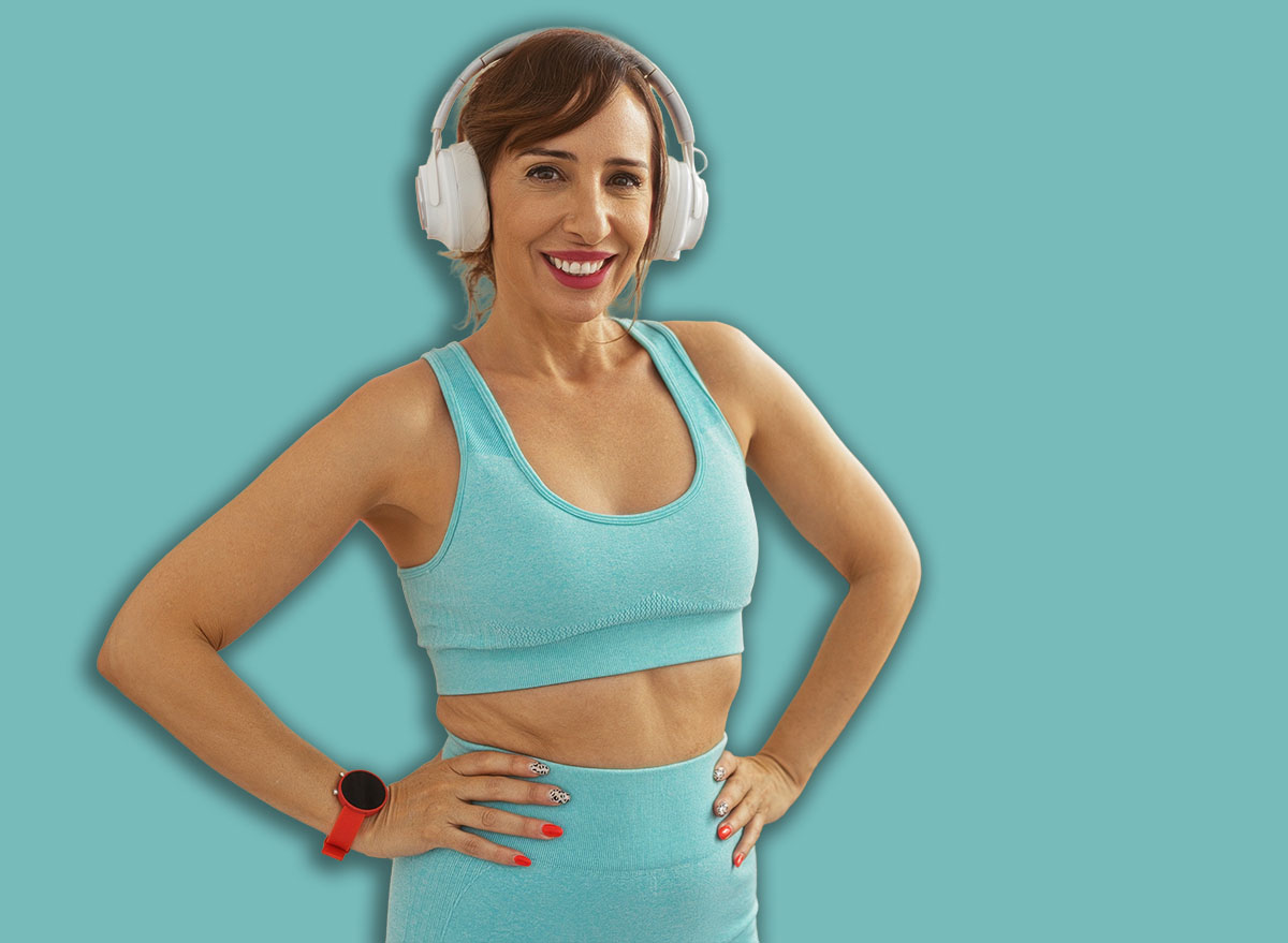 Woman in gym wearing headphones and athletic clothing with hands on hips, smiling confidently in front of yoga mats and exercise balls indoors