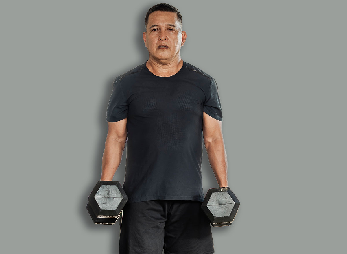 Middle-aged Southeast Asian man carries two dumbbells in a farmer’s walk for forearm or grip strength and endurance training inside open-air gym.